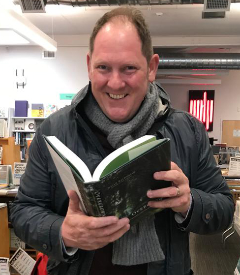 Image of Julian Raxworthy at the MIT Press bookstore holding open his book Overgrown and smiling